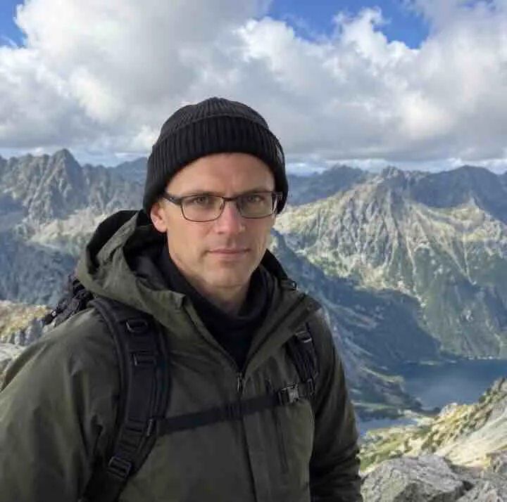 Portrait of Vaclaw Karlfeld in a beanie and glasses, with mountains and a lake in the background.